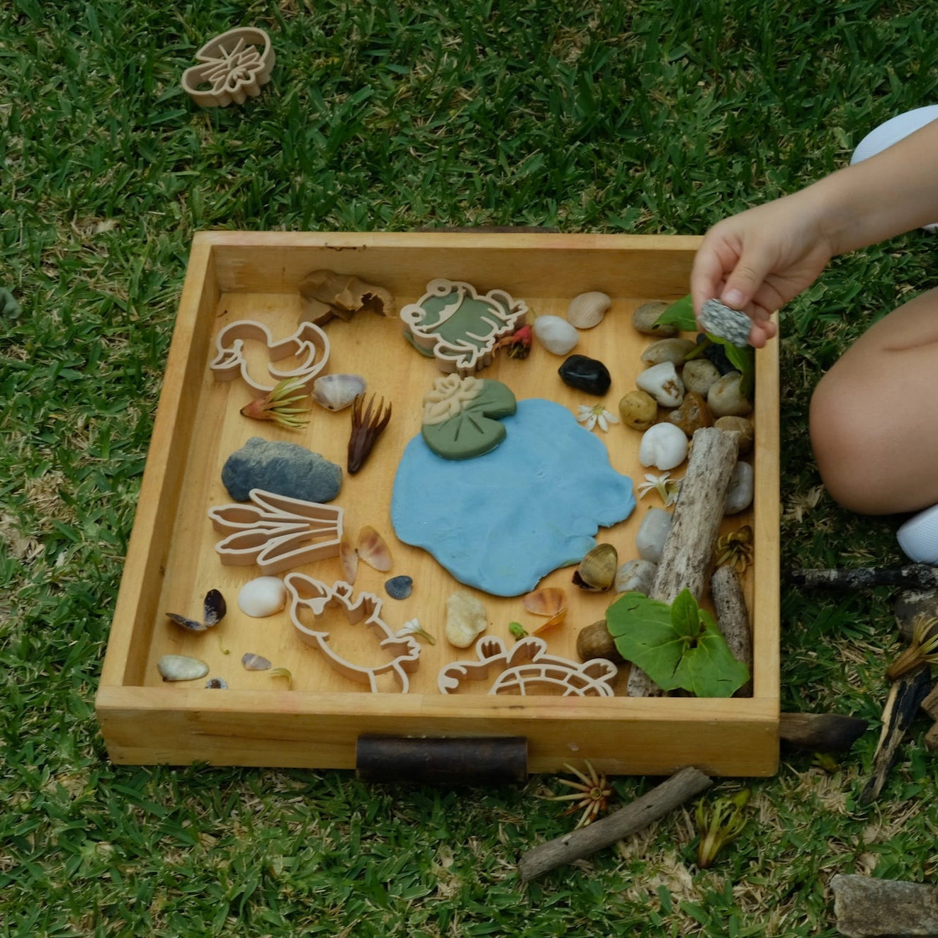 Child playing with a wooden tray filled with natural materials on grass