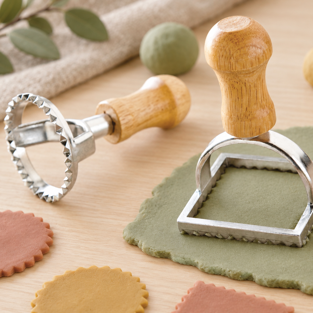A pasta cutter and stamp set with wooden handles and silver metal edges, shown on a light wood surface with rolled dough and colorful cutout pasta pieces.