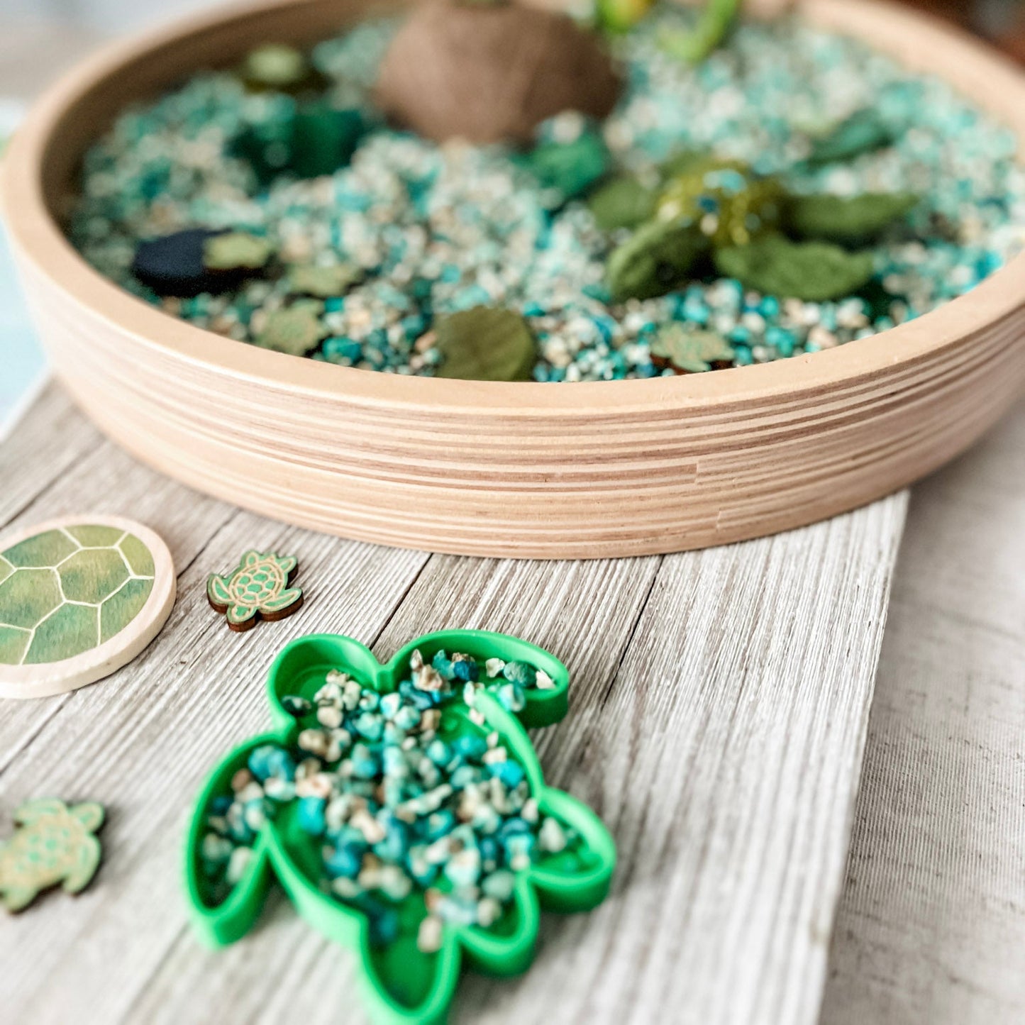 A shallow round wooden tray filled with small green leaves, blue-green pebbles, and a brown rock, styled on a light wood surface with green cookie cutters and shell-like pieces nearby.