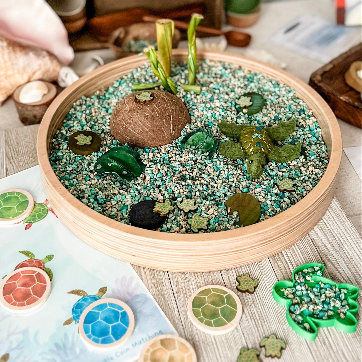 A round wooden tray filled with decorative sea turtle-themed items, including green and blue sand, small shells, a coconut shell, and faux seaweed, displayed on a light wood surface with matching turtle-shaped pieces and a printed turtle card nearby.