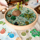 A round wooden tray filled with decorative sea turtle-themed items, including green and blue sand, small shells, a coconut shell, and faux seaweed, displayed on a light wood surface with matching turtle-shaped pieces and a printed turtle card nearby.