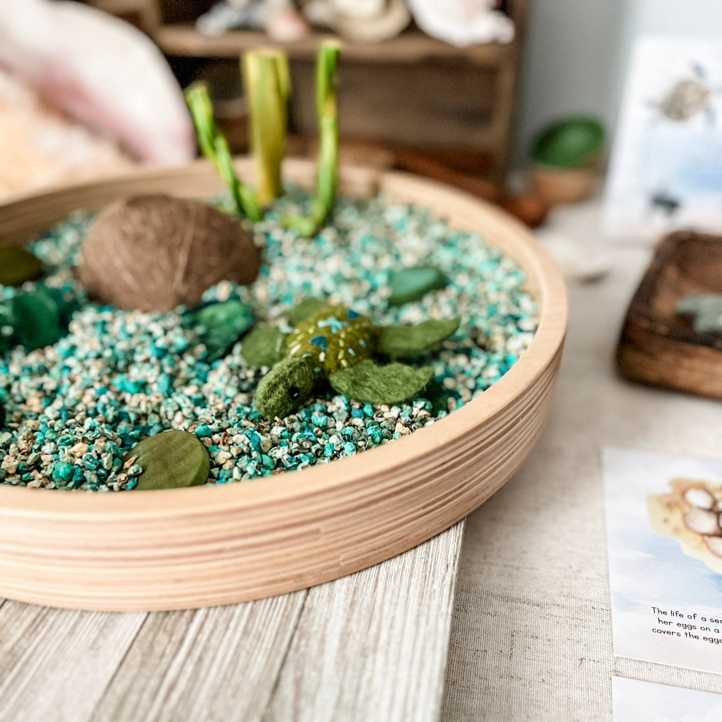 A round wooden tray filled with faux succulent plants and turquoise-colored decorative gravel, displayed on a light wood surface with a blurred indoor background.