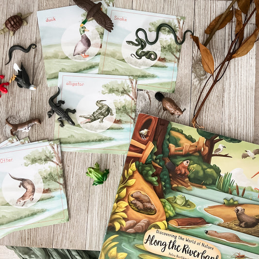 A children's nature-themed book and matching cards laid out on a light wood surface, surrounded by small animal figurines, dried leaves, and branches. The book cover shows a colorful riverbank scene with animals, and the cards show illustrated animals in circular frames.