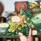 A hand holding open a colorful children's picture book showing an illustrated forest scene with animals and text bubbles. The background is softly blurred.